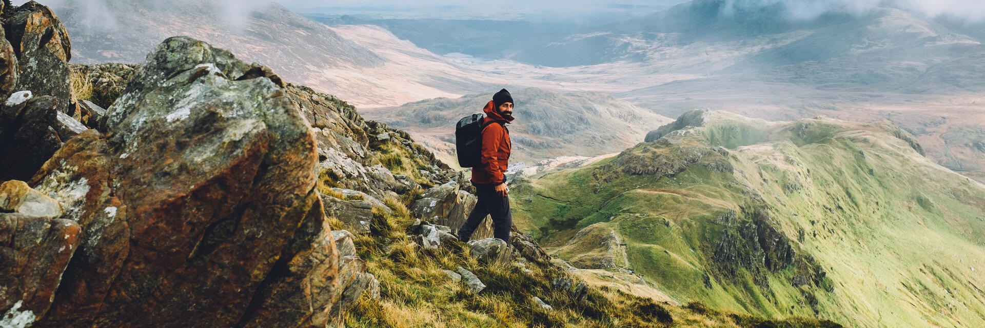 Ein robust gekleideter Mann, der den Gipfel einer großen Bergkette mit Panoramablick erklimmt.