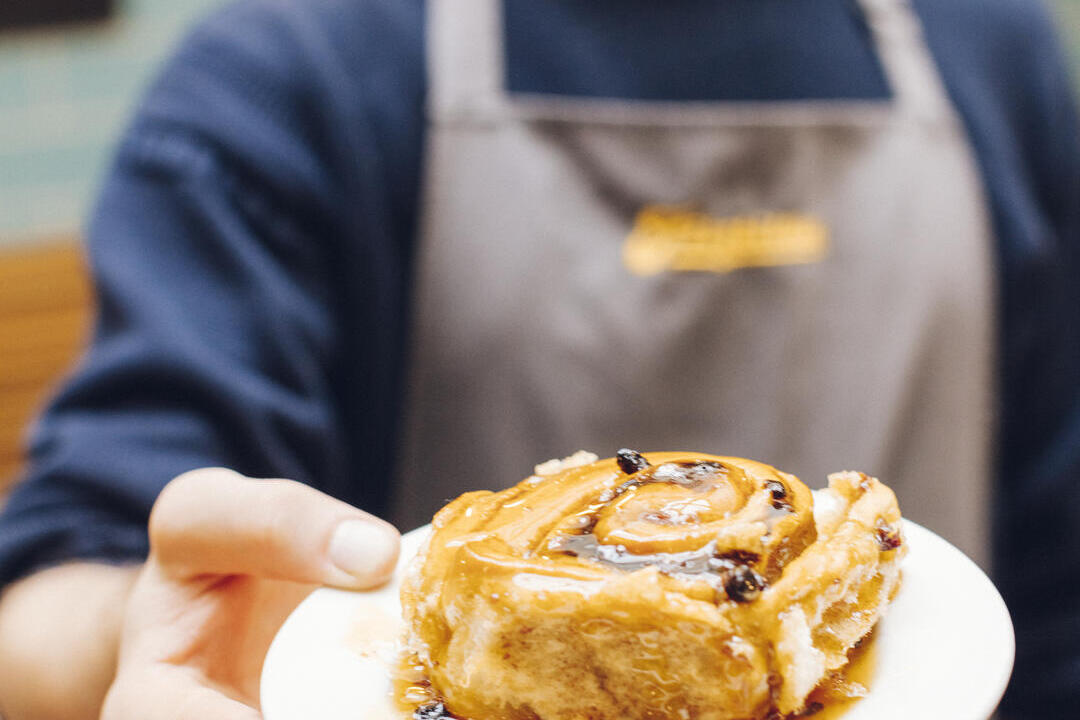 Smiling person wearing an apron holds a plate with a sticky cinnamon roll, focus on the pastry, blurred café background.