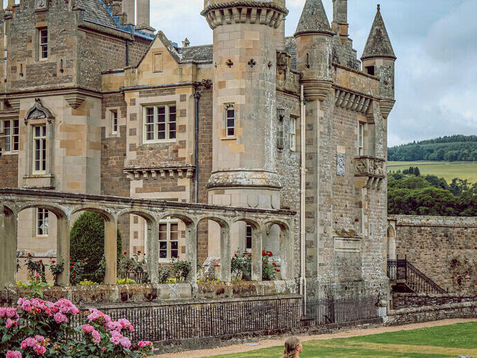 Historic stone castle with turrets and arched walkway, lush gardens, pink flowers, and a woman twirling on the lawn under a cloudy sky.