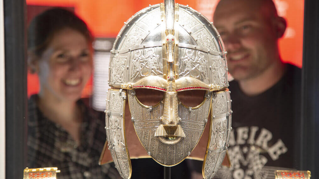 Two people admire an ornate Anglo-Saxon helmet in a museum display with a bright orange backdrop.