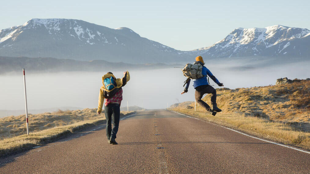 Two men walking and jumping on a road surrounded by mountains