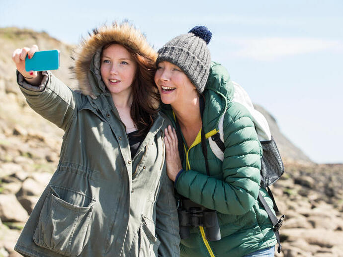 Mutter und Tochter machen ein Selfie an der Küste, sie tragen Hüte und Mäntel