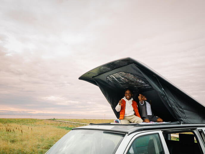 Two children sitting and smiling in a rooftop tent on a car, with grassland and water in the background under a cloudy sky.