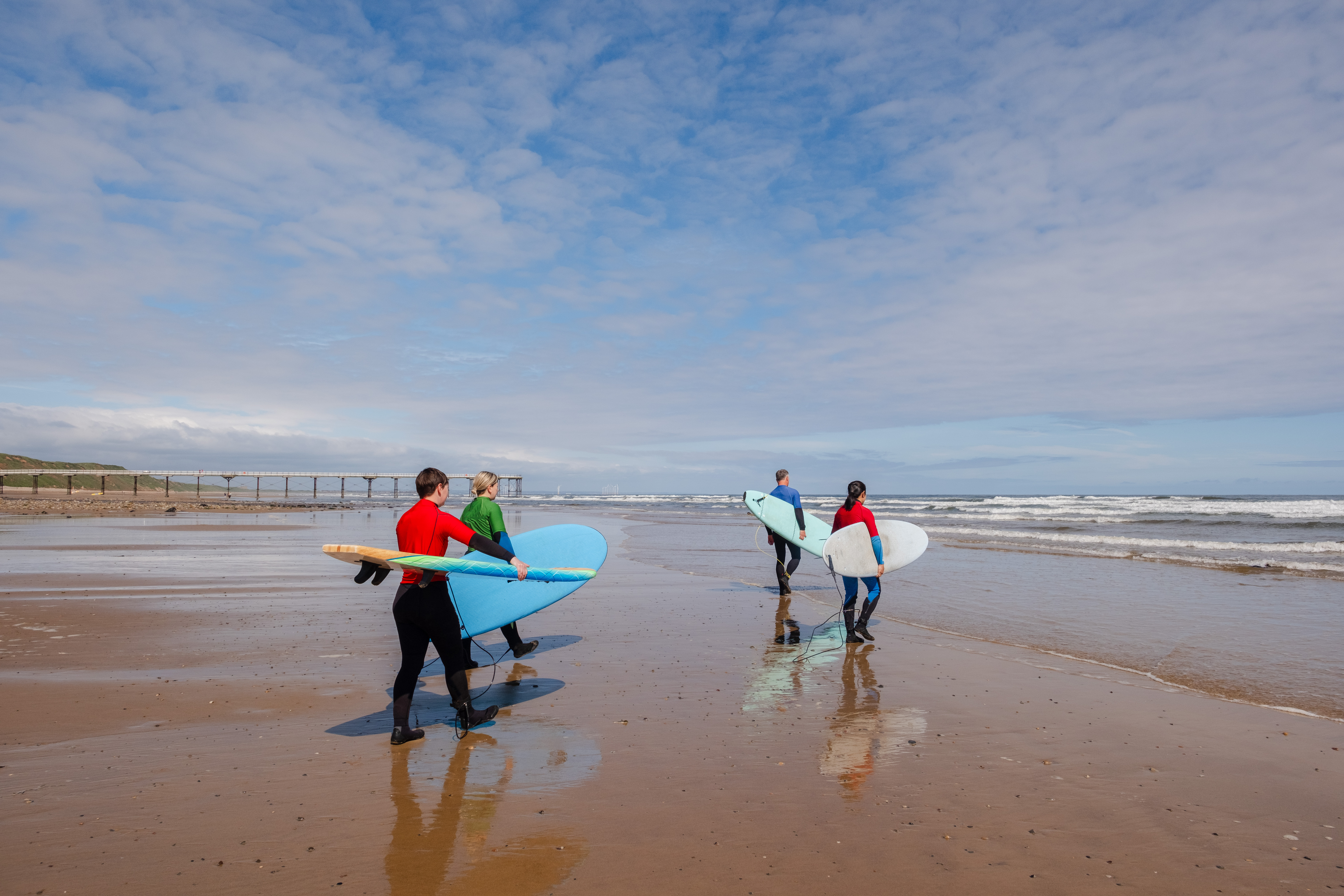 Four people walk toward the sea wearing wetsuits and carrying surf boards
