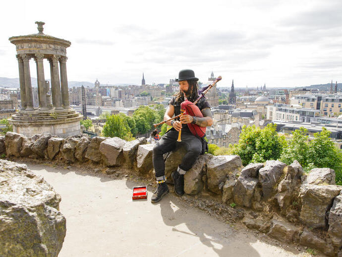 Dudelsackspieler sitzt auf einer Steinmauer mit Stadtblick und klassischem historischen Denkmal links.