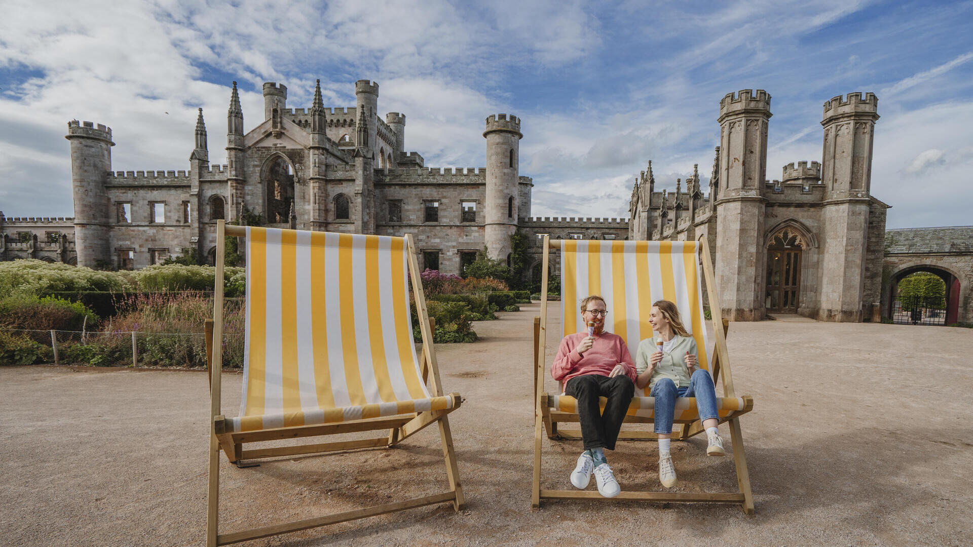 Un homme et une femme sont assis sur de grandes chaises de plage rayées jaunes et blanches devant un château historique, mangeant des glaces.
