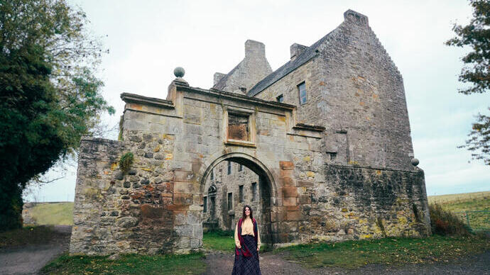 A woman stands in front of the stone entrance of a ruined castle, with grassy ground and trees around.