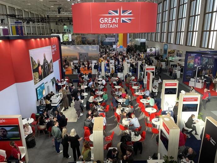 Busy convention hall with Great Britain branding, red and white decor, many attendees seated at tables, large windows, and exhibition booths.