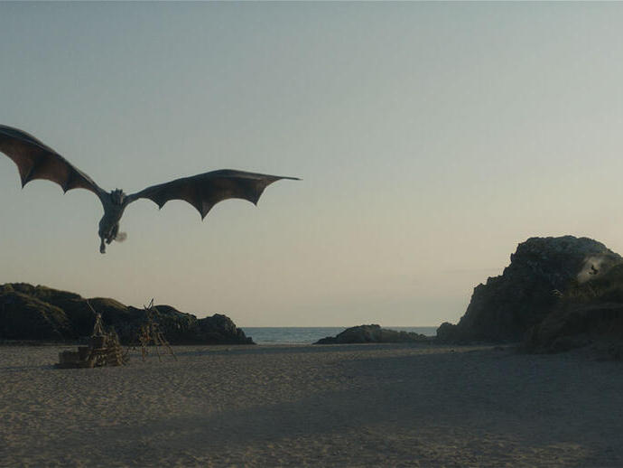 A large dragon flies over a sandy beach with rocky outcrops and the sea in the background, under a clear sky.