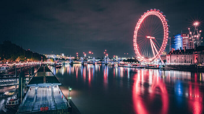La grande roue de Londres illuminée en rouge la nuit, se reflétant sur la Tamise, avec les lumières de la ville en arrière-plan.