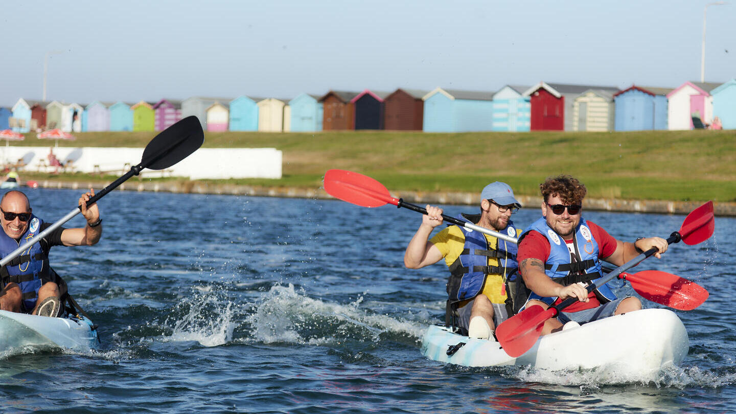 Amigos practicando kayak en la isla de Mersea con cabañas de playa al fondo