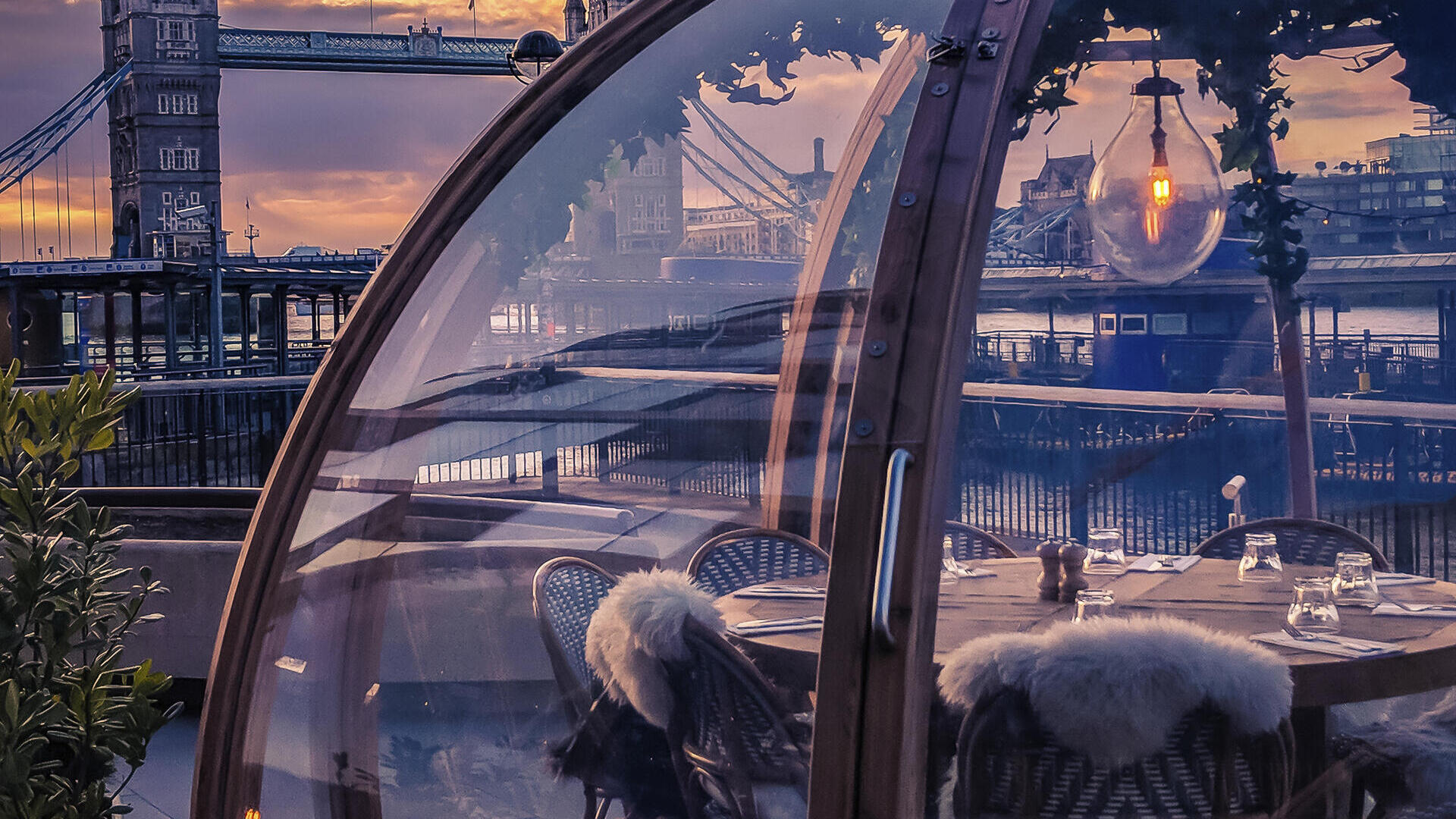 A glass dome enclosing a restaurant table beside a river in the middle of a city