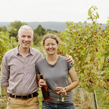 Couple standing in a vineyard holding wine and glasses.