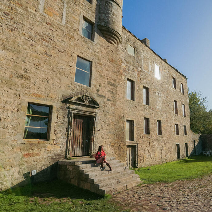 Historic stone building with arched entry gate, green lawn, and cobbled path. A person sits on steps in afternoon sunlight.