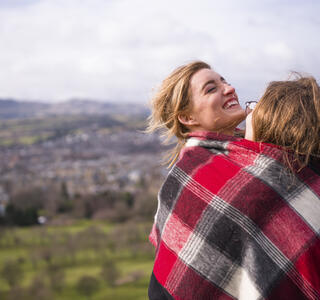 Two women wrapped in red checkered blanket standing on a hill overlooking a city