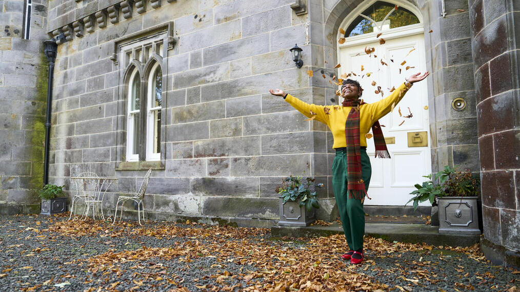 Person stands joyfully in autumn leaves outside a historic stone building with arched windows and door.