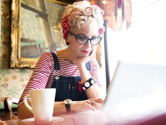 Close up of a blond woman wearing a stripy top and red head scarf, having a coffee and looking down at a lap top screen, smiling.