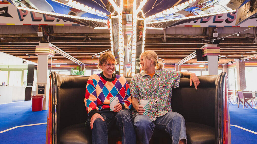 A man and a woman wearing roller skates sit in a retro teacup at a roller disco