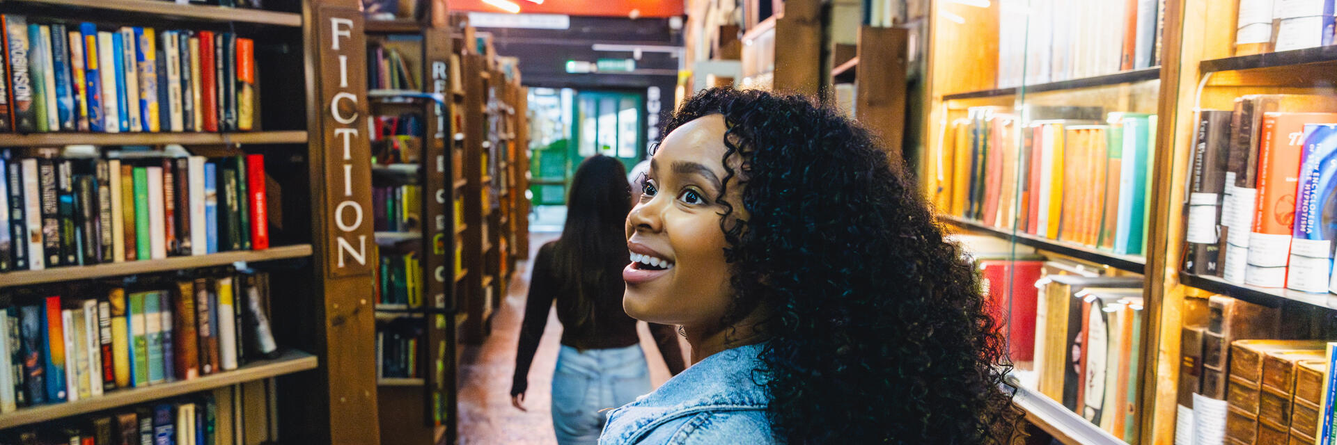 Two people walk through a vibrant, well-lit bookstore with shelves of books, stone walls, and neon lights overhead.