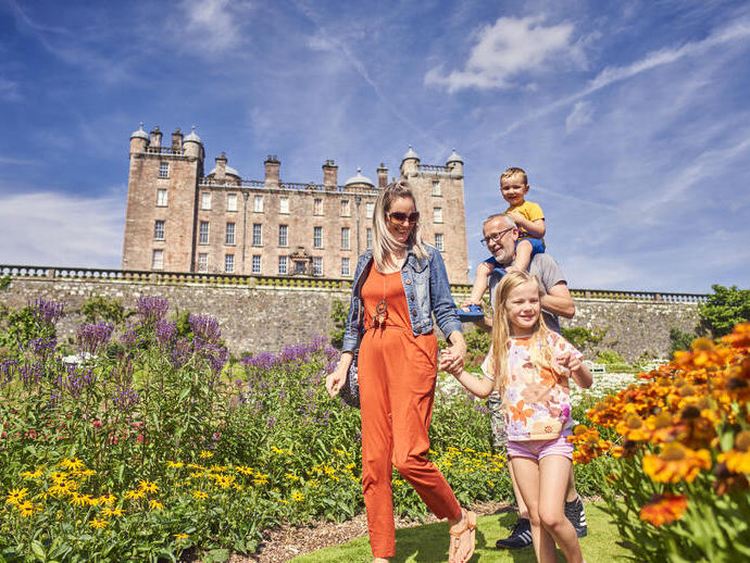 Family walking through colorful gardens in front of a historic stone castle under a blue sky, enjoying a sunny day outdoors.