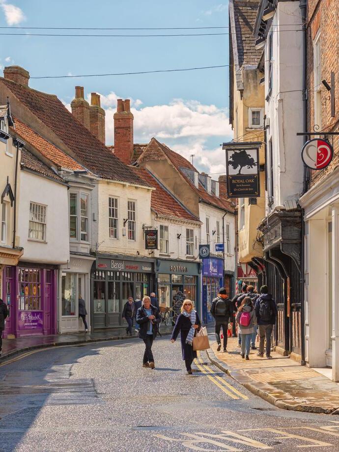 Shopping street just after a rain shower. Historic buildings line the street and shoppers walk between them. A sky with cloud is overhead.