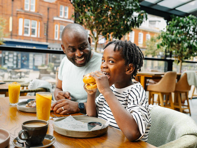 Child eating a burger and adult smiling at an outdoor restaurant table with drinks, plants, and red brick buildings in the background.