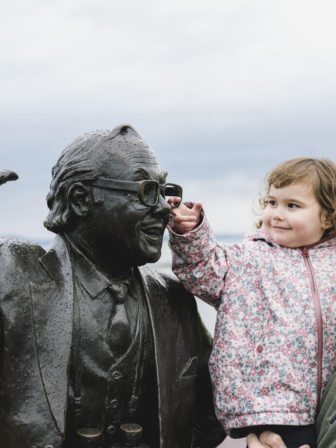 A young child and man stand next to a statue on the seafront, as the child reaches up to touch the statue’s face