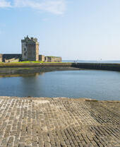 Historic stone castle with tower beside a calm waterfront and stone-paved area, under a clear blue sky.