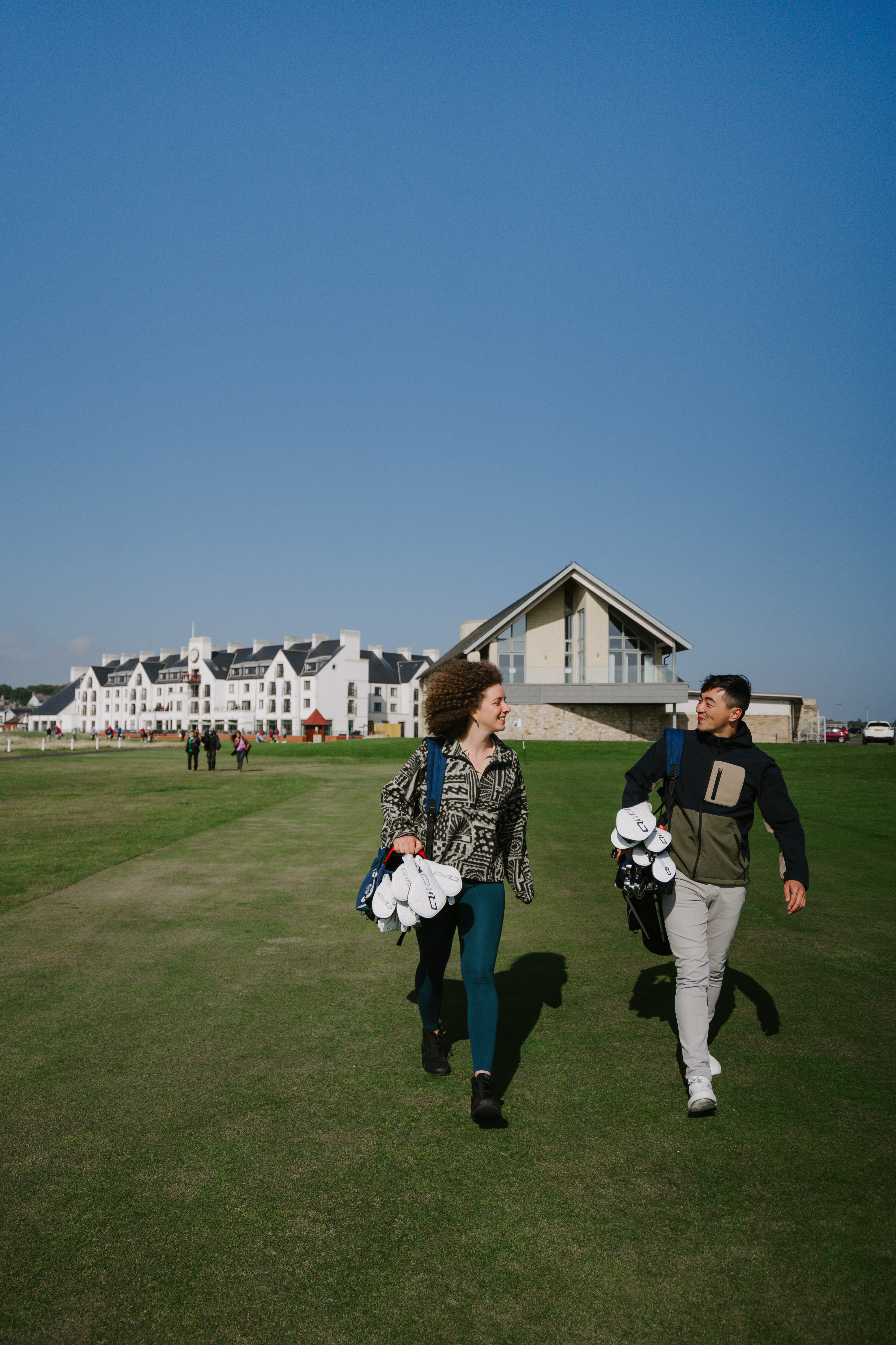 A man and a woman carrying golf equipment walking on the grounds of a country golf club.