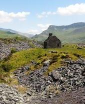 Ruined stone building on grassy hill, surrounded by slate and scattered rocks, with mountains and blue sky in the background.