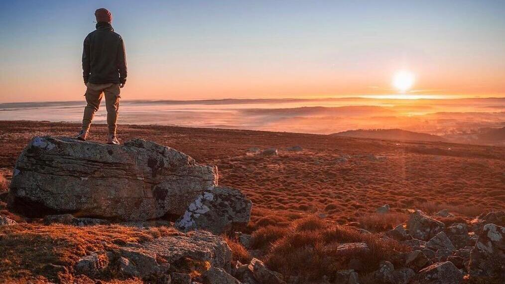 Vista trasera de un hombre de pie sobre una roca, contemplando el campo al amanecer