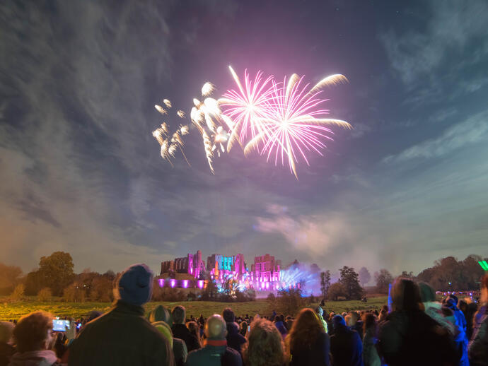 Fireworks display over a ruined castle watched by a crowd of people in the evening
