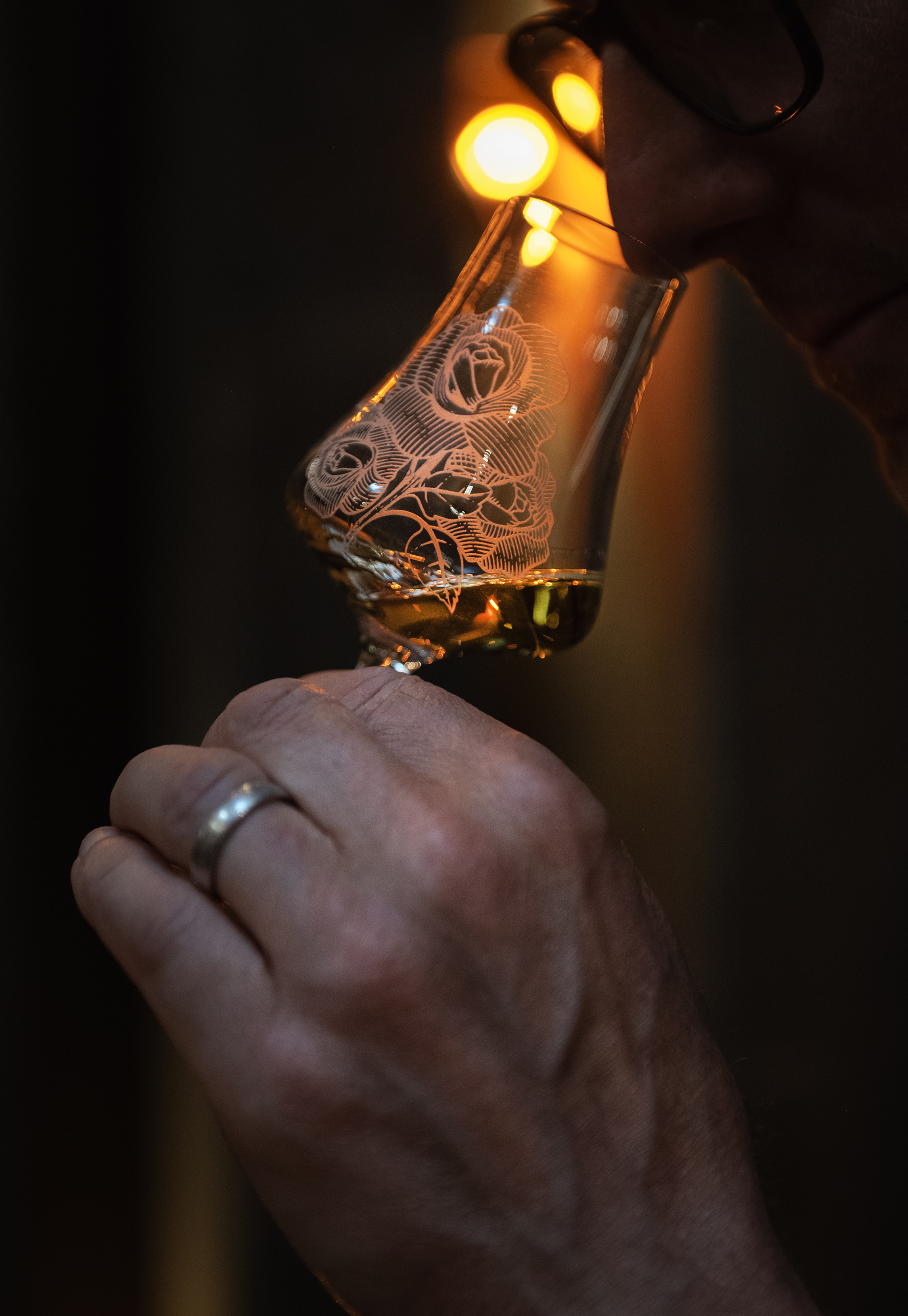 A person smelling a patterned whisky glass with amber liquid, highlighted by warm light in a dark setting, evoking tasting or sensory evaluation.