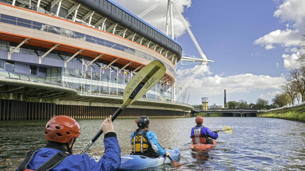 Kayakers passing a stadium along a river.