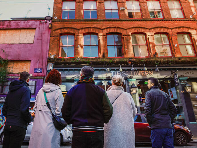 Five people stand on a street facing a red-brick pub called Tib Street Tavern in an urban area with colorful buildings.
