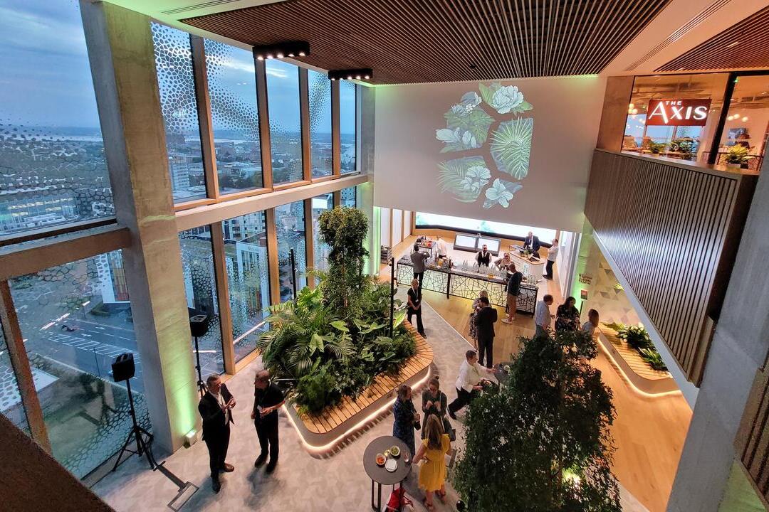 Modern indoor event space with large windows, lush indoor plants, bar area, and people socializing. Urban cityscape visible outside.