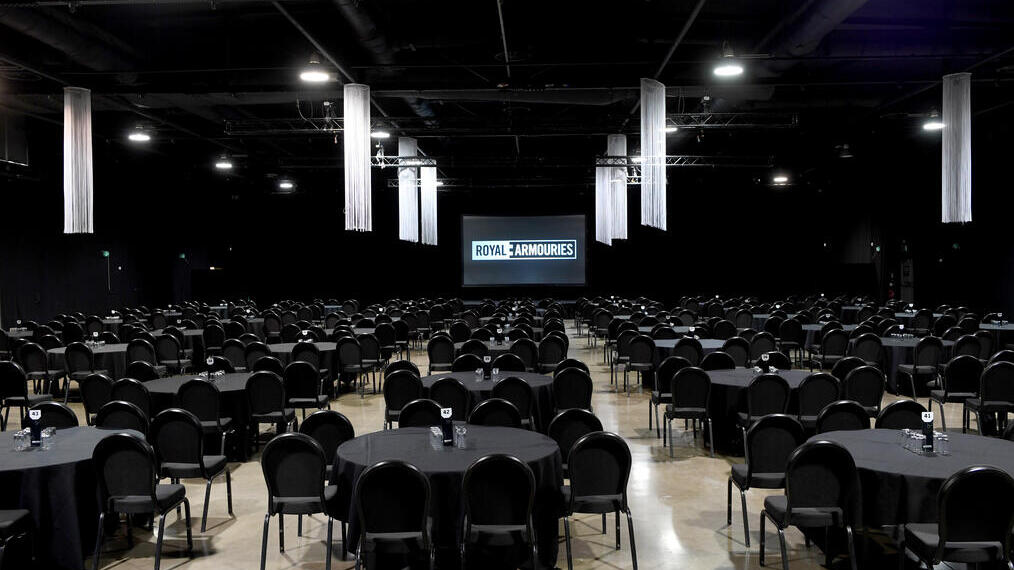 Large banquet hall with round tables and black chairs arranged for an event, stage with screen reading 'Royal Armouries' at the front.