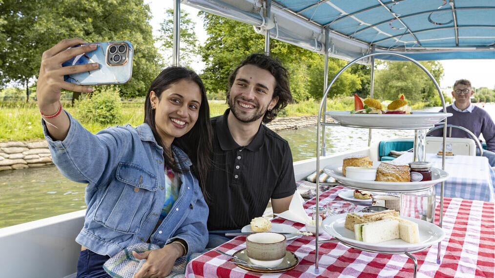 Un couple prend un selfie lors d'un afternoon tea sur un bateau, avec des assiettes de sandwiches et de pâtisseries.