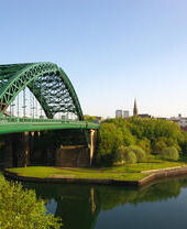 Blick auf eine grüne Eisenbrücke über einen Fluss, umgeben von Bäumen und Gebäuden