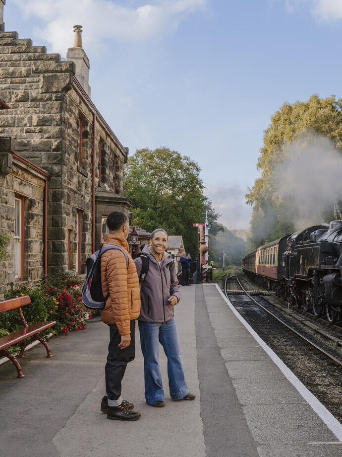 Two people stand talking on a train station platform beside an old stone building with a steam train on the tracks. Trees with autumn foliage are visible in the background.