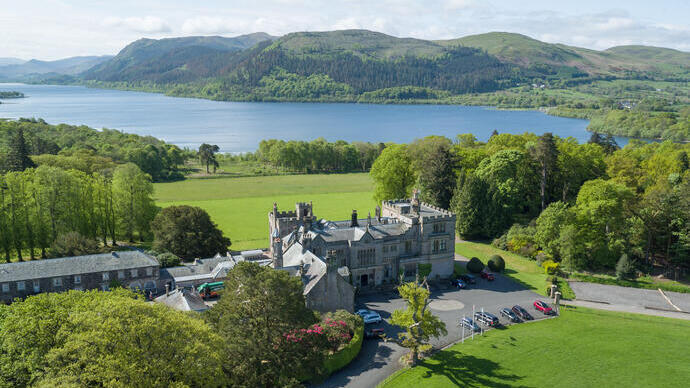 A large historic stone building surrounded by lush greenery, near a lake with hills in the background under a blue sky.