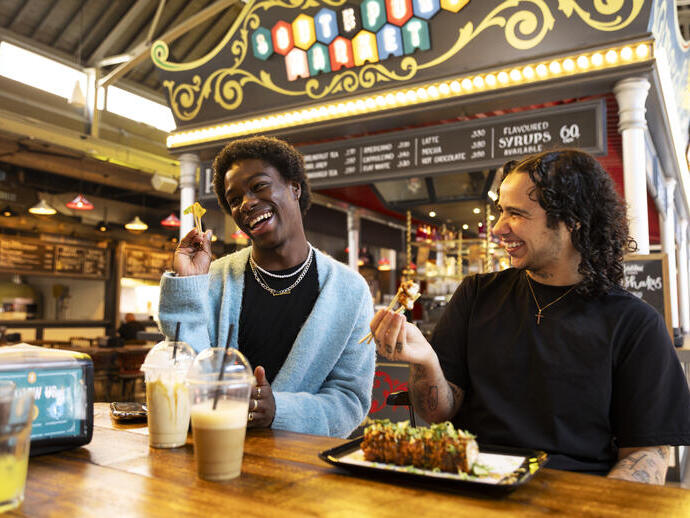 Two friends laugh while eating and drinking inside a food market