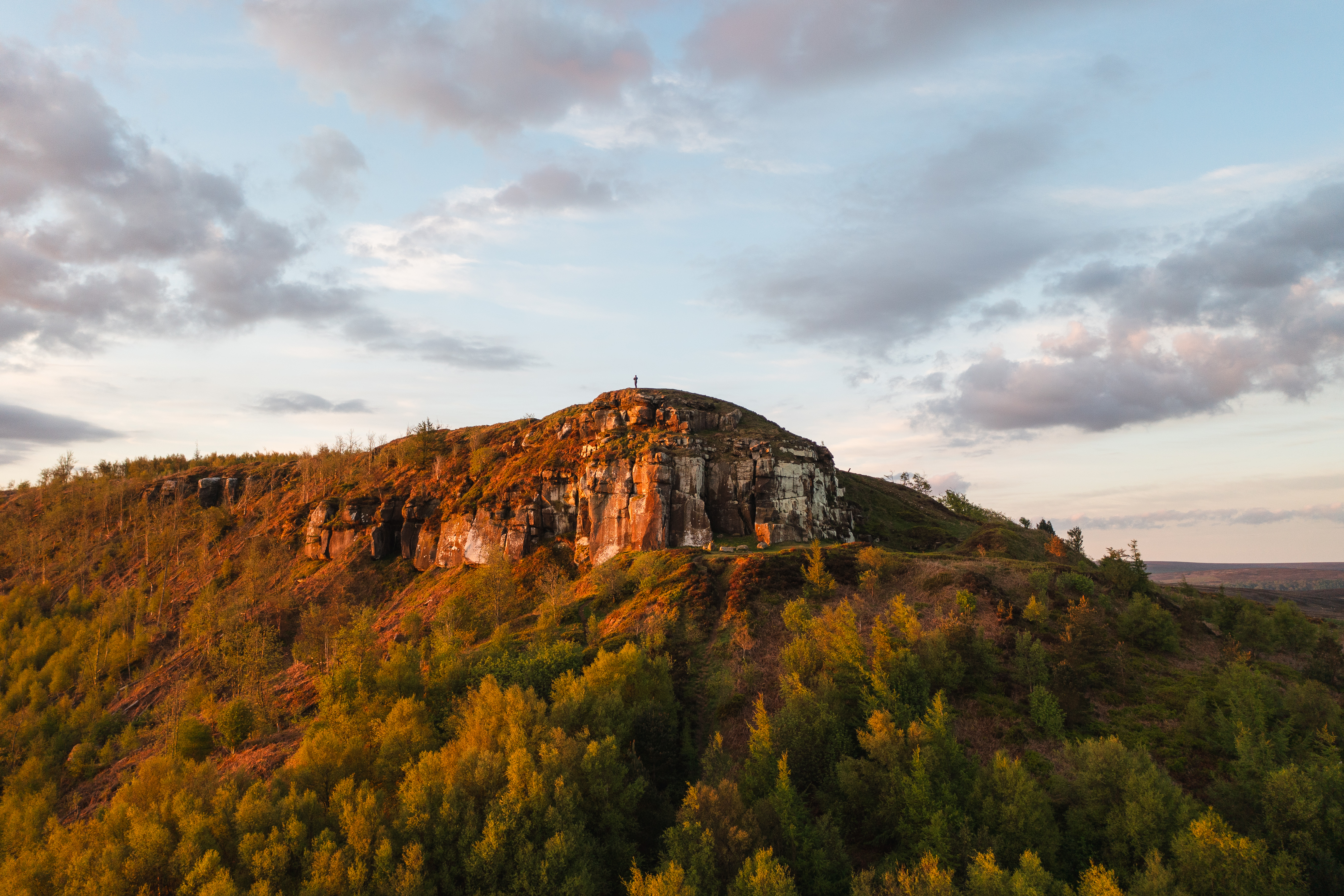 A lone person stands on a cliff top with forest below