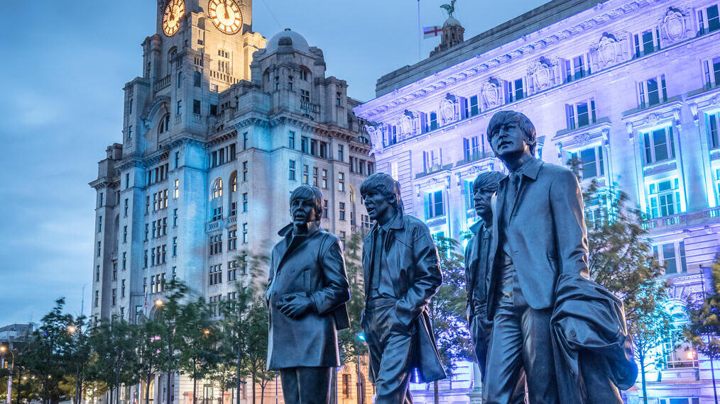 A sculpture of four men with a large building with clock tower behind, lit up in the evening.