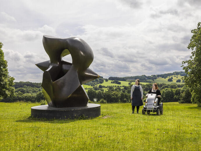 Two women look at a large sculpture set in green gardens with landscape beyond