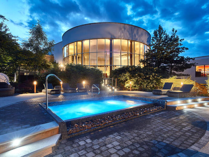 The outdoor spa area at dusk with a lit outdoor spa pool surrounded by loungers and the glass fronted circular spa building in the background. 