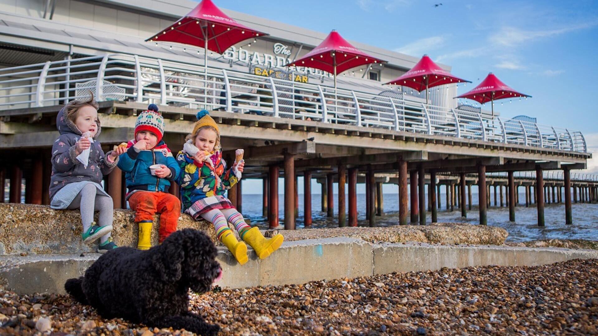 Three children eat ice cream near a pebble beach, with a black dog in front and a pier with red umbrellas in the background.