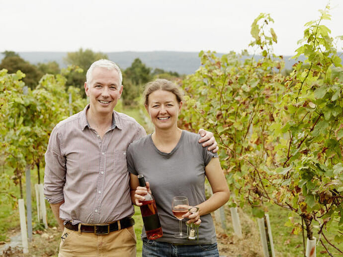 Couple standing in a vineyard holding wine and glasses.