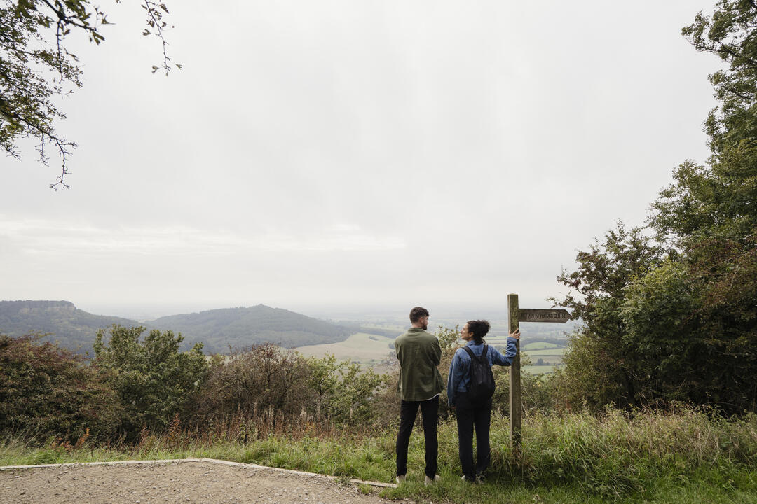Two people stand at a viewpoint with a signpost, overlooking a scenic valley with rolling hills and cloudy sky, surrounded by trees and greenery.
