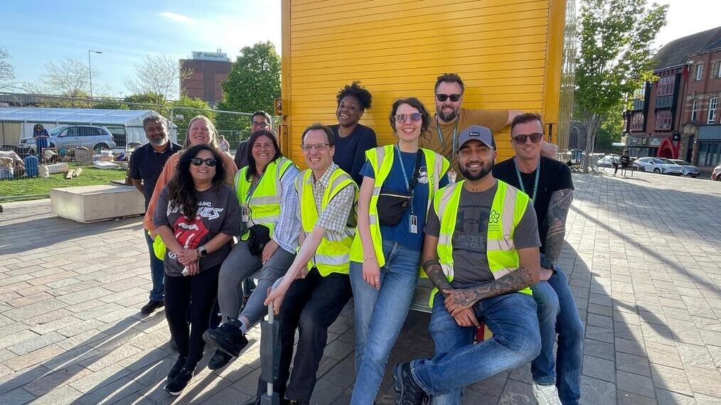 A group of people wearing high-visibility vests and casual clothes pose and smile together outdoors in front of a yellow truck on a sunny day.